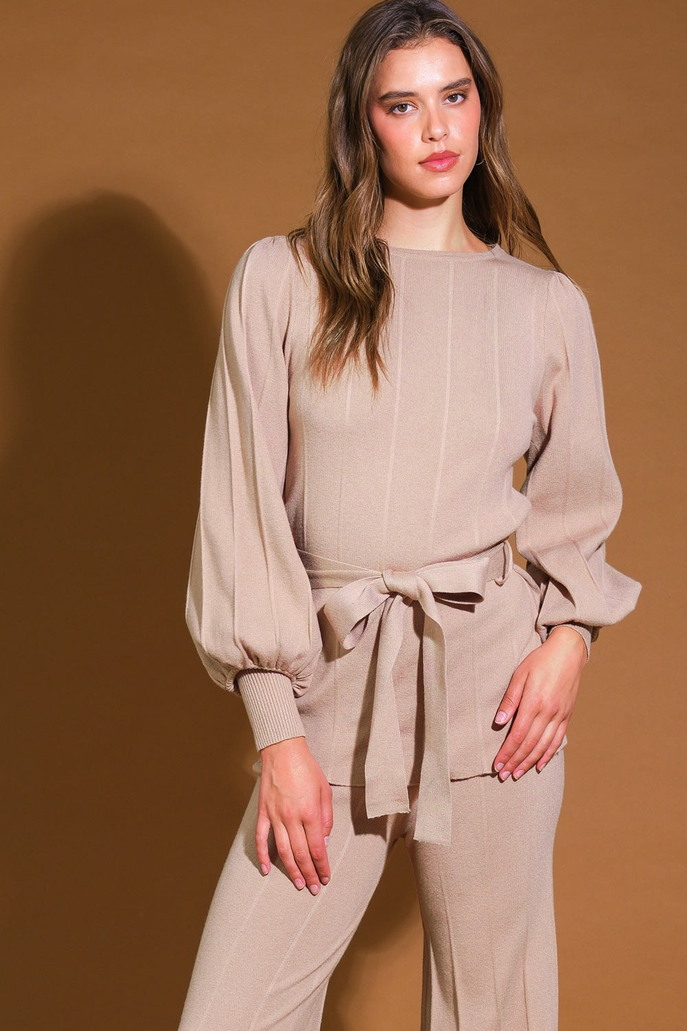 Woman wearing a beige two-piece lounge set with cropped top and jogger pants, standing by a cozy couch.